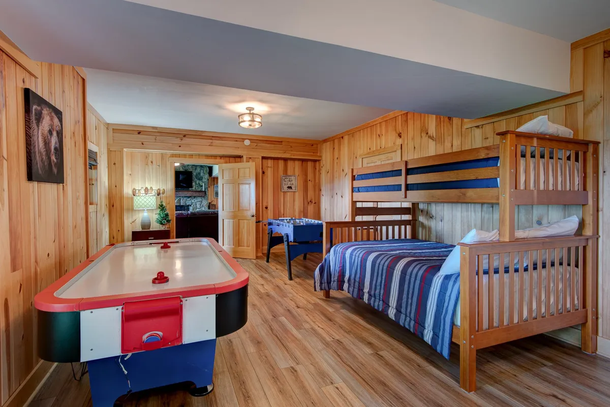 A fun and vibrant room with an air hockey table in the foreground, cozy bunk beds with striped bedding to the side, wood-paneled walls, and playful bear artwork. The space radiates energy and is tailored for game nights and memorable family sleepovers.