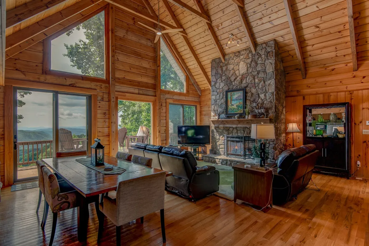 This spacious, open-concept living area features a large stone fireplace, expansive windows with mountain views, a wooden dining table for eight, and comfortable sofas. The vaulted, exposed-beam ceiling contributes to the sense of space and grandeur, creating a welcoming environment for family meals and cozy evenings.