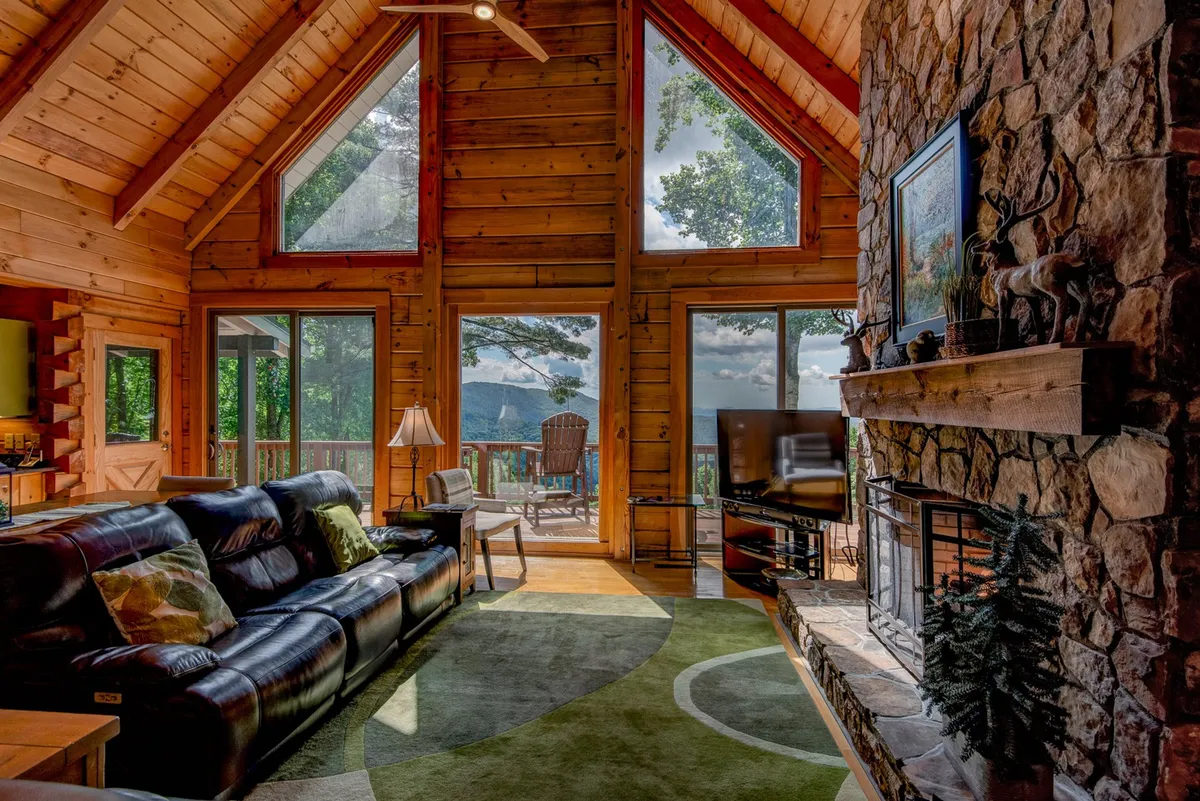 A warmly lit great room featuring a large stone fireplace as the focal point. Expansive floor-to-ceiling windows showcase breathtaking Blue Ridge mountain views. A plush leather sofa and colorful area rug provide inviting seating, creating a cozy atmosphere ideal for relaxing, entertaining, or family gatherings.