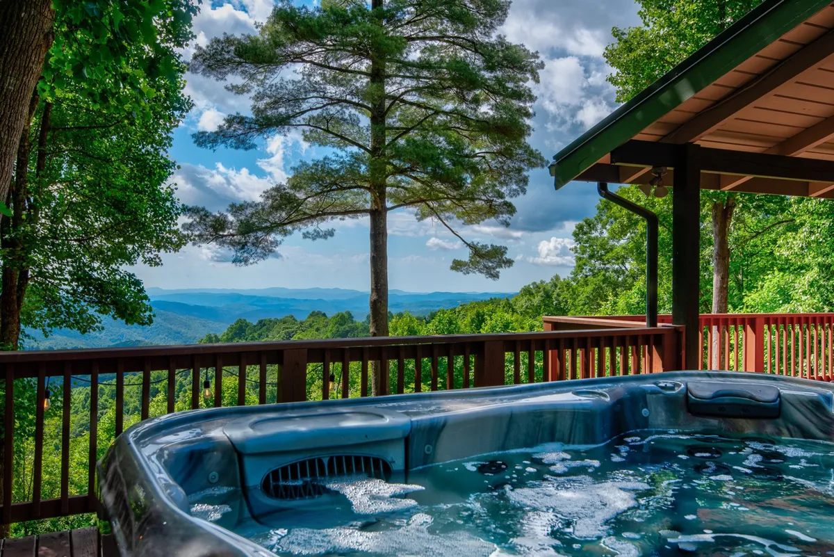 The image showcases the exterior deck of a mountain cabin nestled among towering trees, with a steaming hot tub in the foreground. Beyond the deck, breathtaking layered mountain ridges stretch into the distance, bathed in natural light. The scene radiates tranquility, inviting guests to soak in the hot tub while savoring unobstructed panoramic views or gathering with friends as the sun sets.