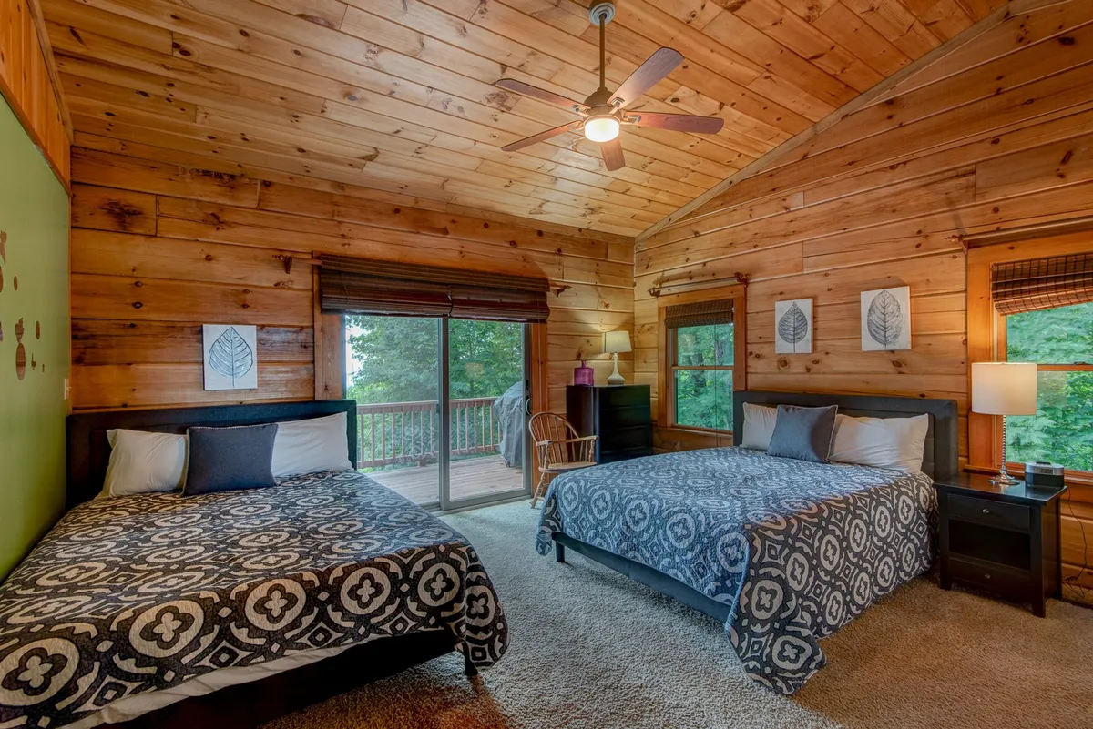 A comfortable bedroom with two beds, a wooden ceiling, and a sliding door opening to a balcony with nature views. The decor is relaxed and inviting, enhanced by natural light and tasteful wall art.
