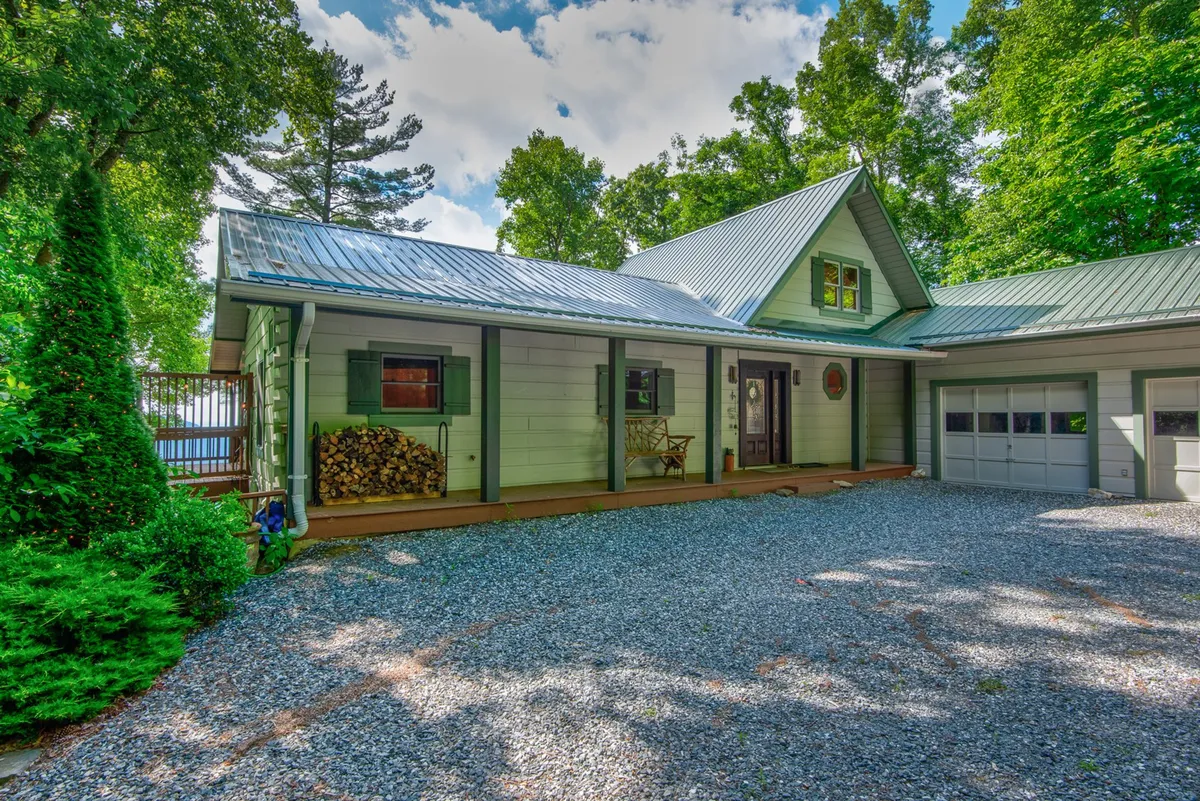 The front of the mountain retreat, featuring a welcoming porch with green shutters and a gravel driveway. A woodpile sits nearby, and guests are seen relaxing on the porch, while others gather around a fire pit ready for an outdoor cookout. The mood is sociable and inviting, showcasing both the home’s curb appeal and its outdoor gathering spaces.