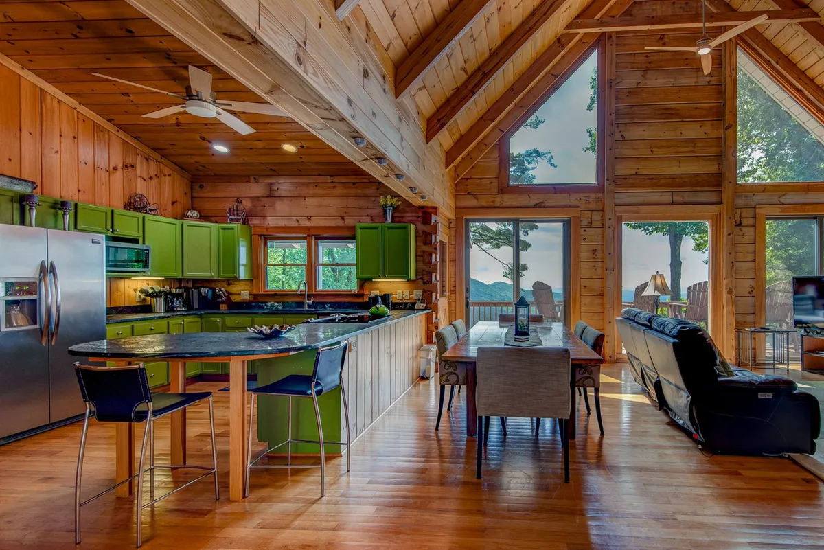 A bright, fully equipped kitchen with distinctive green cabinetry, a large island with bar seating, and generous windows showcasing exterior views. The adjoining dining table sits nearby, making it a hub for both meal prep and shared family moments.