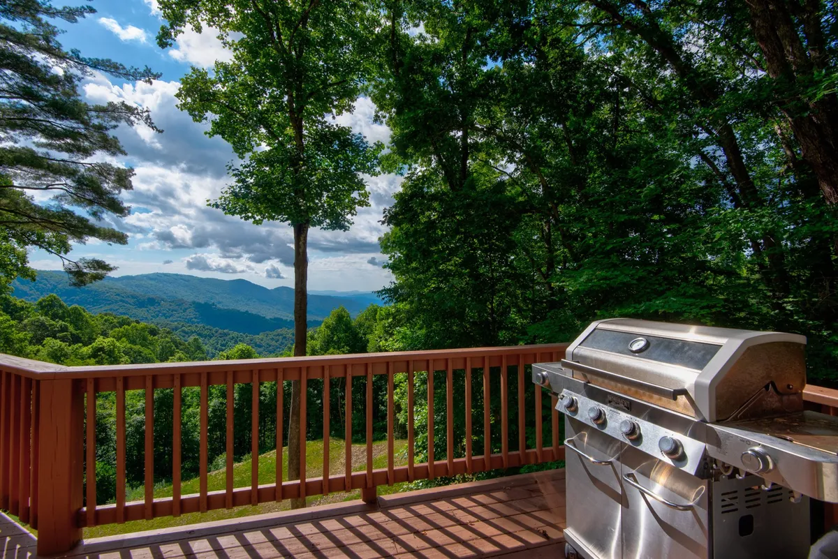 A tranquil deck scene with a grill in the corner, classic Adirondack chairs lined up along the railing, and a wide-open mountain view stretching into the distance. The golden evening light bathes the deck, hinting at sunset, while the setup invites friends or couples to relax and socialize outdoors. The setting feels both private and panoramic, ideal for sharing drinks or a barbecue under the stars.