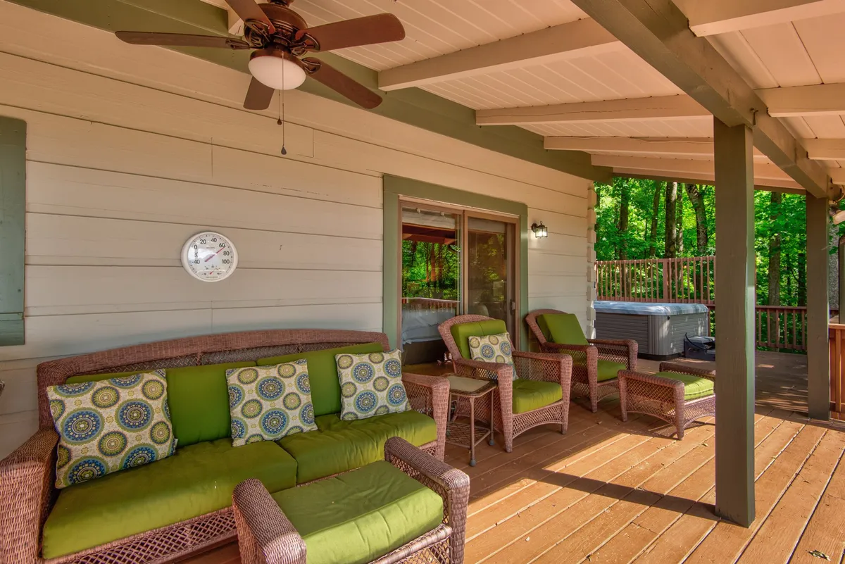 A comfortable wooden deck featuring woven seating with green cushions and a hot tub nestled at the edge, surrounded by lush greenery. The view from the deck extends into the trees beyond, creating a tranquil outdoor living area for relaxation and socializing.