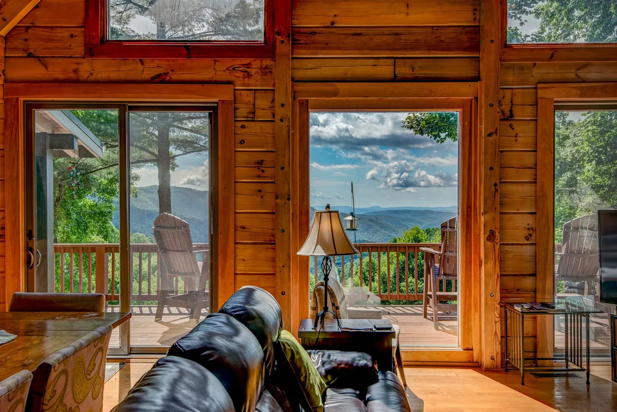 The photo showcases a cozy living area featuring a comfortable black leather sofa, large windows that frame breathtaking mountain views, a wooden dining table, and soft lighting from a decorative lamp. The space exudes warmth and is filled with natural light, creating an inspiring atmosphere for work and relaxation.