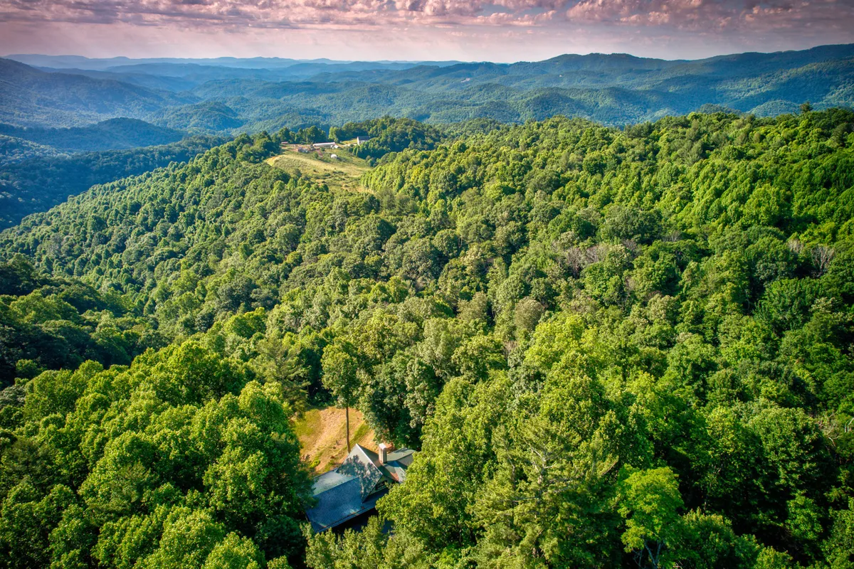 This sweeping view captures lush green forests, rolling hills, and a distant mountain range, with a single, isolated residence visible in the expanse. The landscape radiates peace, seclusion, and inspiration from the surrounding wilderness.